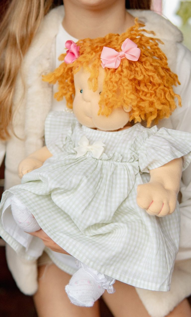 muñeca de tela personalizada unica baby doll Niña con cabello rizado y muñeca vestida con un vestido claro y cintas rosas.