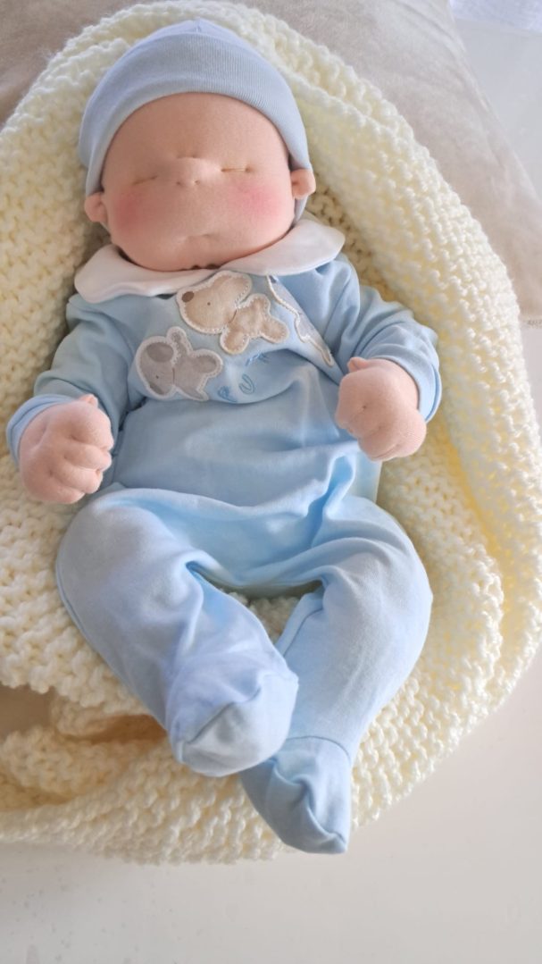 Bebé de tela con gorro azul y pijama celeste con un osito bordado