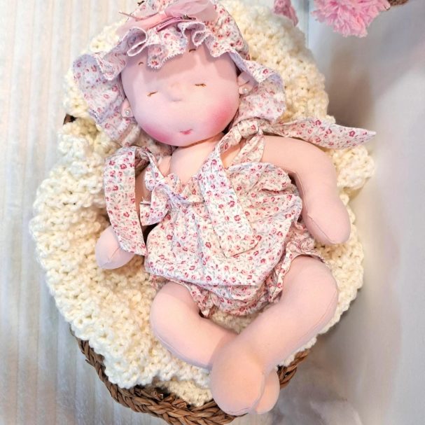 Bebé de tela con gorro y pelele de flores rosas, sobre una manta de punto crema
